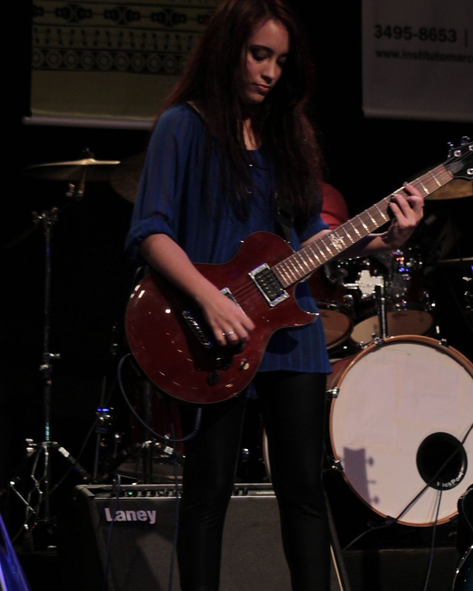 Kalina tocando guitarra vermelha no palco, com bateria e amplificador ao fundo.